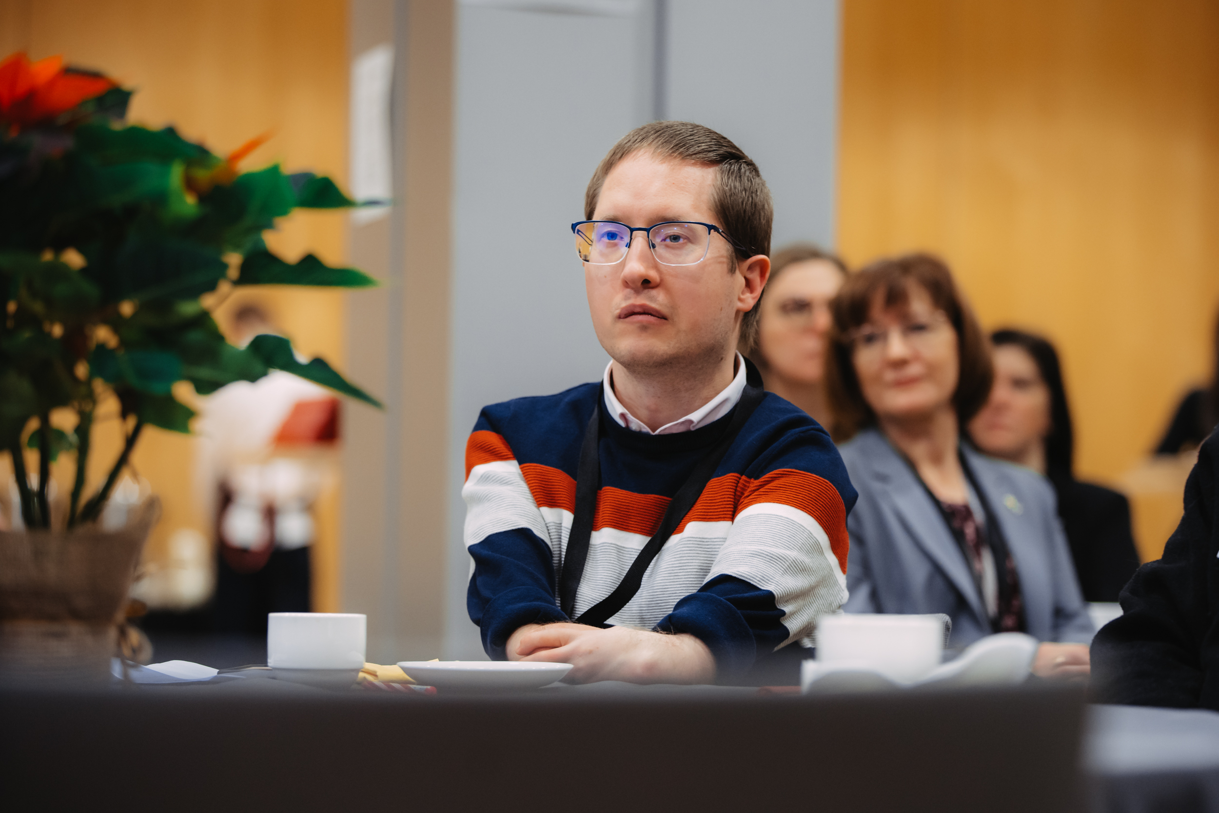 Konferences dalībnieki, priekšplānā jauns vīrietis svītrainā džemperī un ar brillēm
