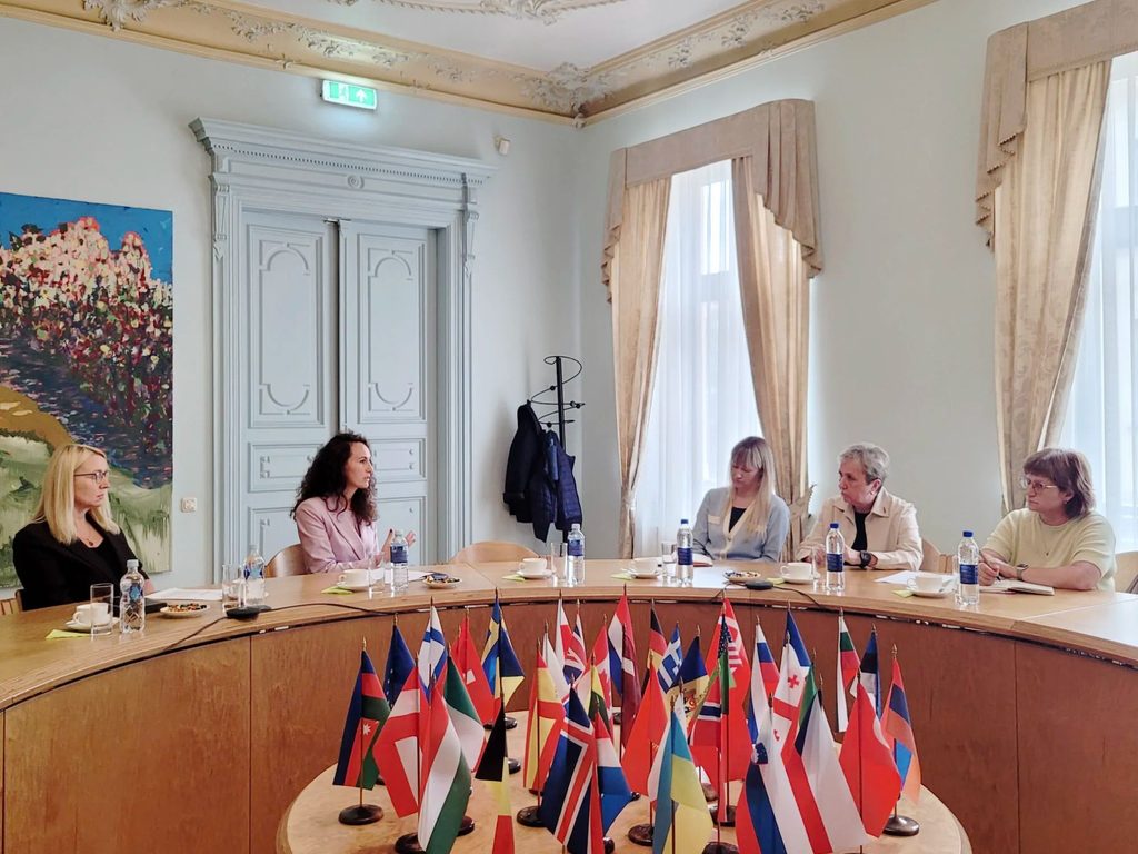 Sešas sievietes sēž pie liela koka galda formālā telpā. Viena sieviete runā, žestikulējot ar rokām, kamēr citas klausās. Galda centrā izvietoti daudzi mazi dažādu valstu karodziņi.