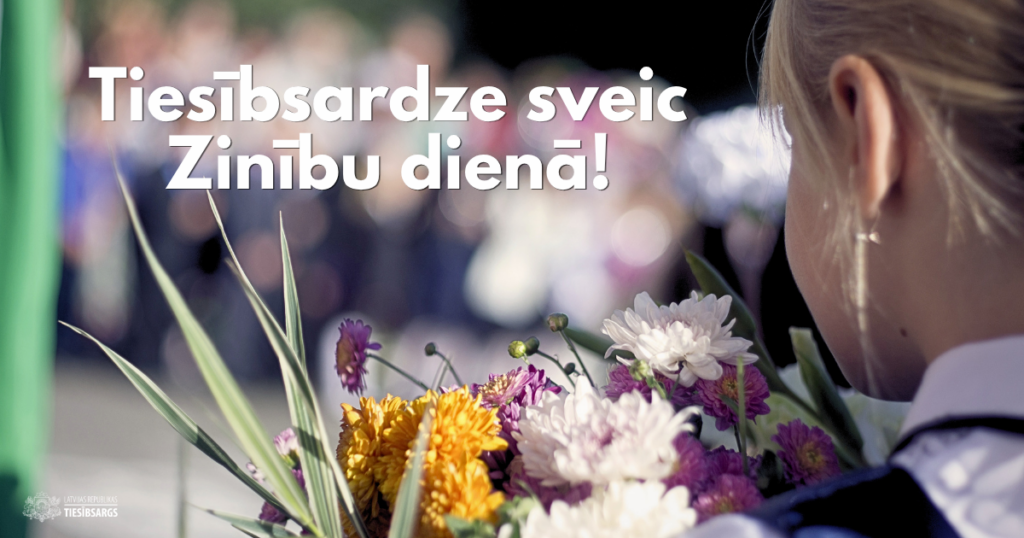 meitene no mugurpuses ar blondiem matiem tur krāsainu ziedu pušķi. Attēlā redzams teksts: "Tiesībsardze sveic Zinību dienā!". Fons ir izplūdis.