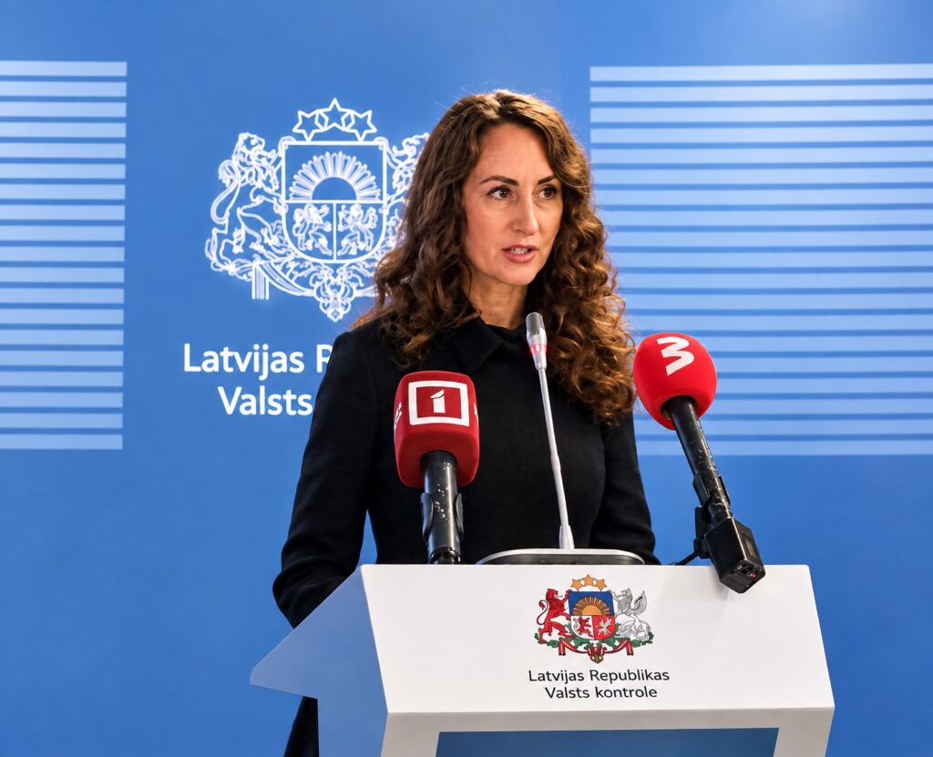 Latvijas Republikas Tiesibsardze Karina Palkova. Uz pults redzams Latvijas ģerbonis un teksts "Latvijas Republikas Valsts kontrole". Priekšā ir trīs mikrofoni, tostarp sarkani ar "1" un "3" logotipiem. Fonā zila siena ar Latvijas ģerboni un daļēji redzamu tekstu "Latvijas F Valsts".