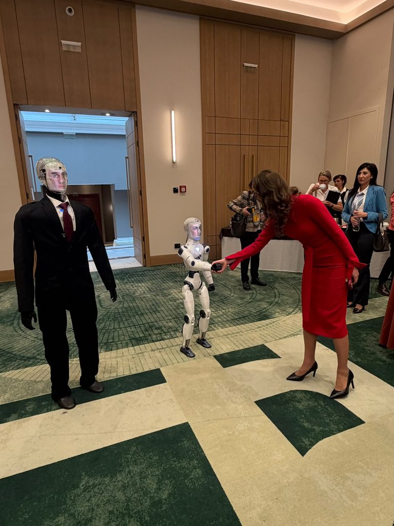 Sieviete sarkanā kleitā sniedz roku mazam, baltam robotam. Liels robots melnā uzvalkā stāv kreisajā pusē, bet fonā redzami citi cilvēki pasākuma telpā ar koka sienām un zaļi bēšu rakstainu paklāju.