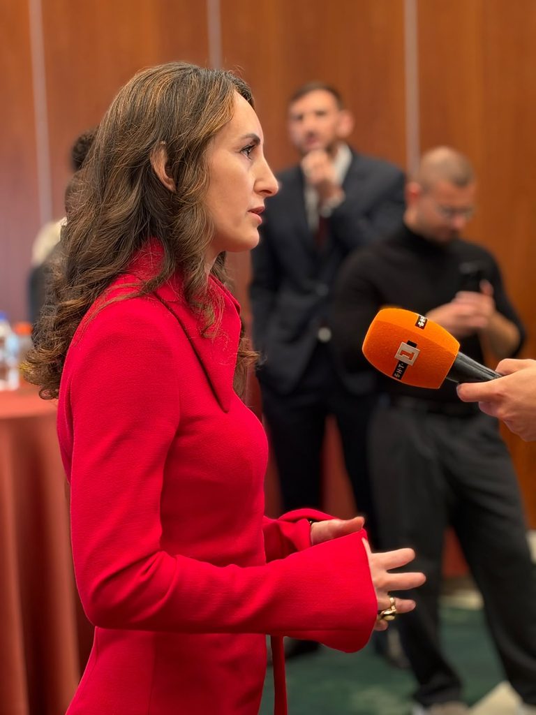 Sieviete sarkanā kleitā ar viļņotiem brūniem matiem sniedz interviju oranžam mikrofonam. Fonā izplūduši cilvēki.